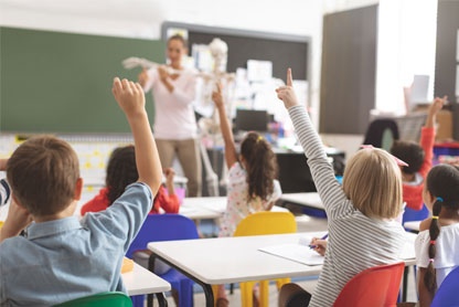 Classe con all'interno una maestra che spiega e i bambini che alzano le mani